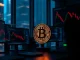Bitcoin coin on a trading desk with multiple monitors showing crypto price charts and market data.