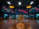 Bitcoin coin on a table in a dim trading floor with monitors showing financial charts