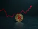 Bitcoin coin on dark surface with faint red downward trend chart in background, representing bearish market risk.