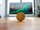 Bitcoin coin on table with monitor showing price chart in background