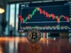 Bitcoin coin on desk with financial chart on monitor in background