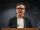 Andy Burnham speaking at a press conference with digital blockchain background