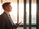 Professional businessman in suit standing in modern office with city skyline, representing financial executive